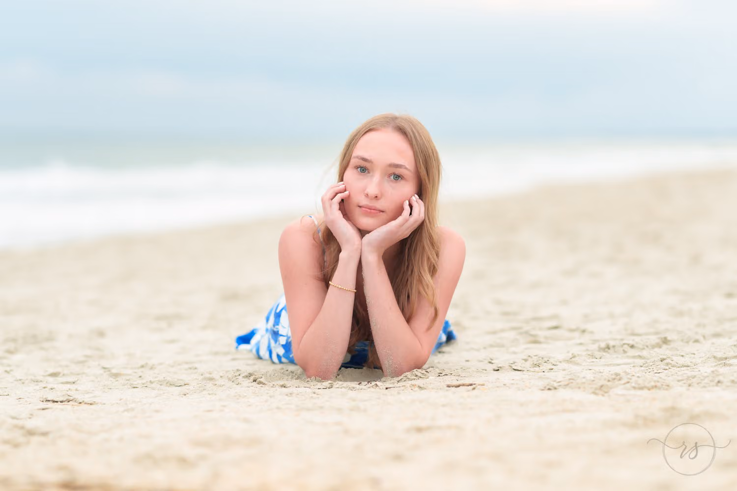 High school senior in a blue dress lying in the sand with her chin in her hands at Huntington Beach State Park