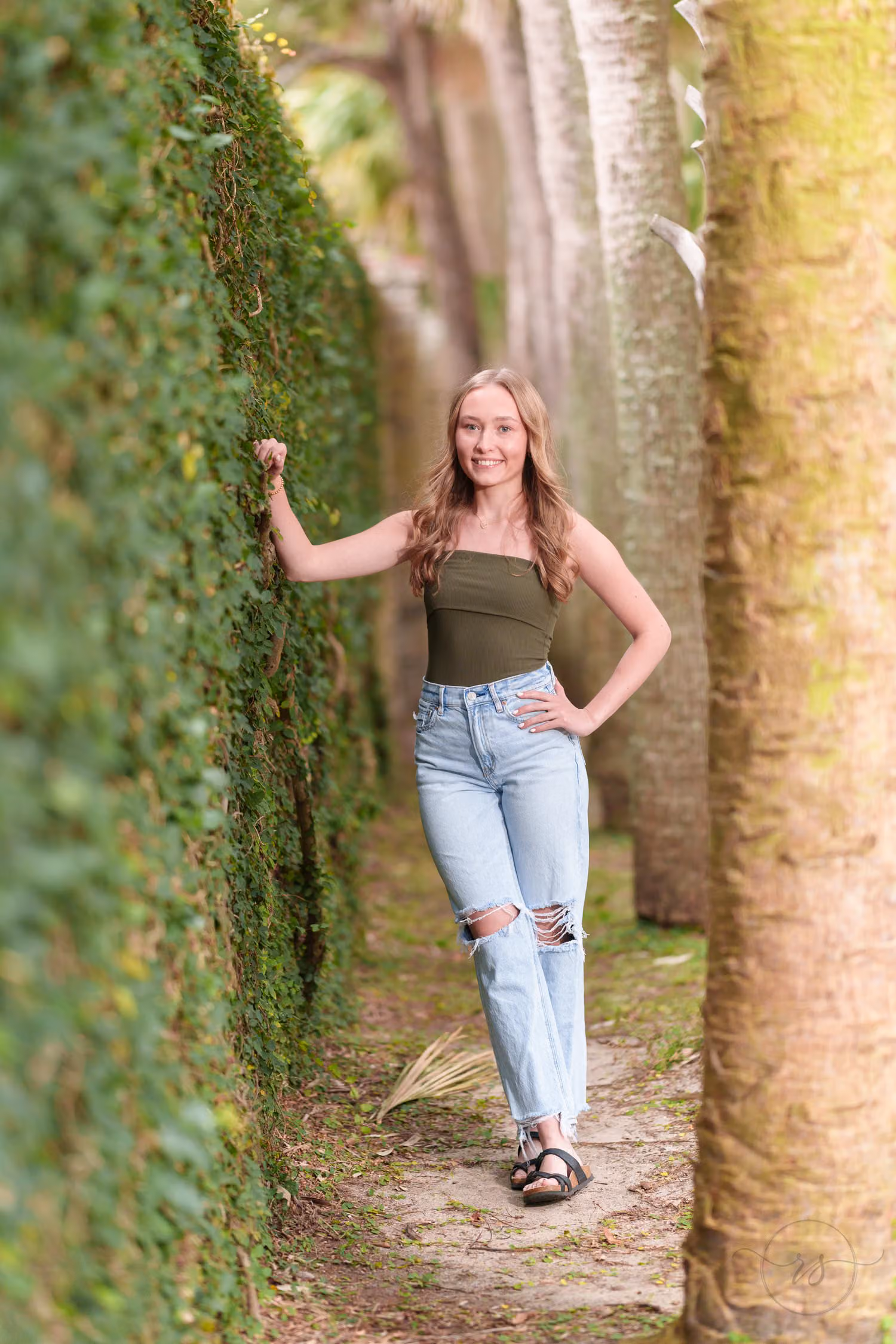 High school senior in jeans and an olive top leaning on the ivy-covered wall by the palms at Huntington Beach State Park