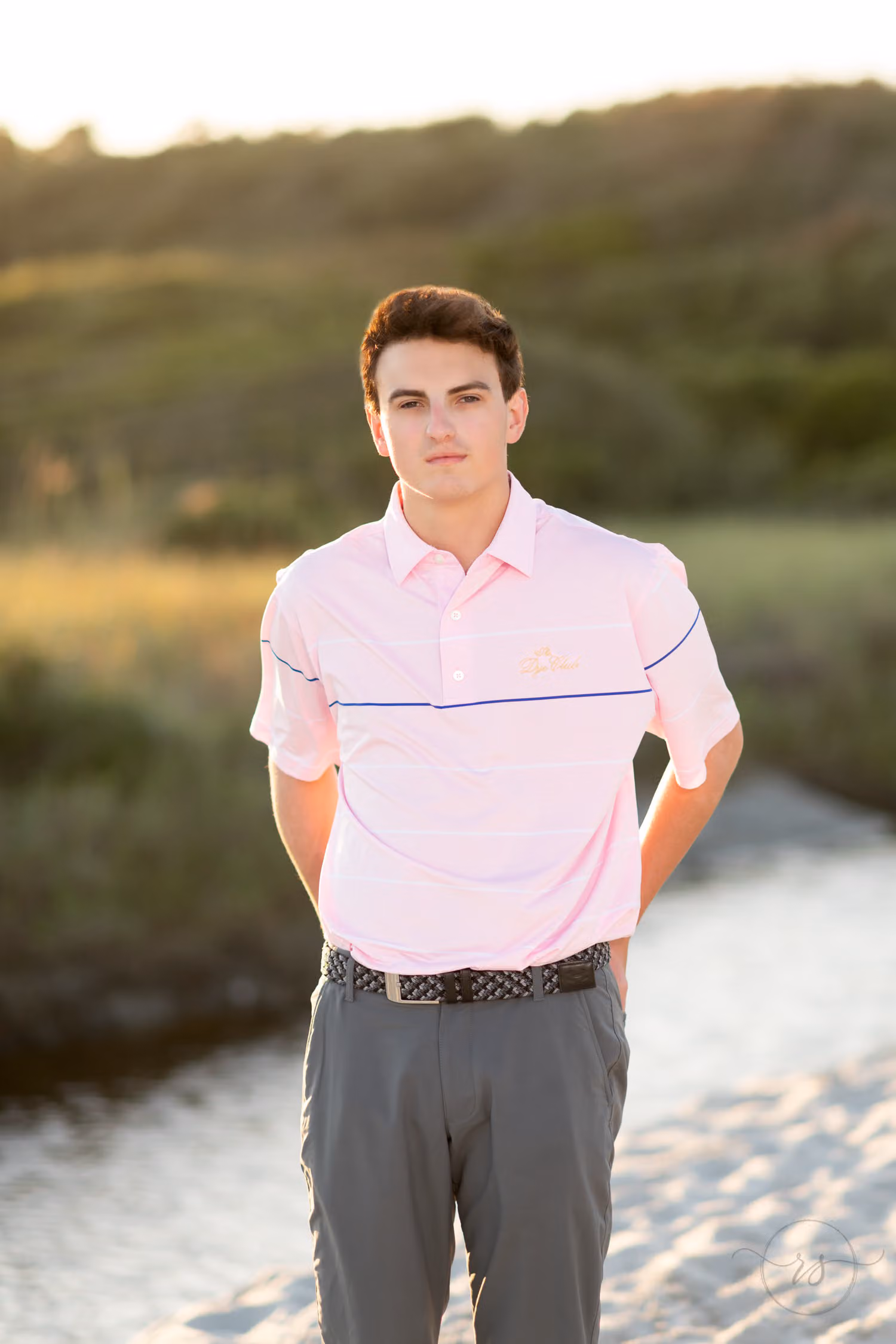 Senior guy in a pink polo standing by a beach creek and dunes in warm sunset light