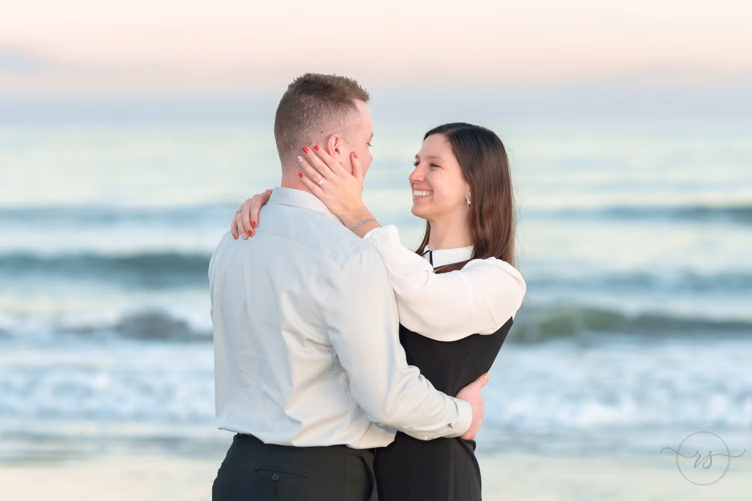 Woman smiling as her fiancé holds her face by the ocean in pastel evening light at Huntington Beach State Park