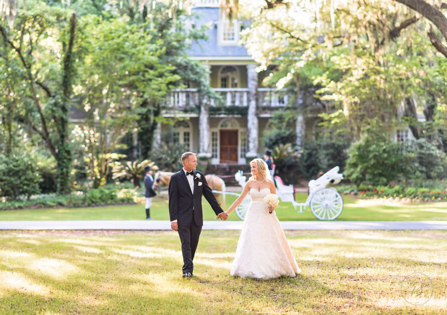 Bride and groom holding hands on the lawn in front of a white carriage and the historic house at Wachesaw Plantation