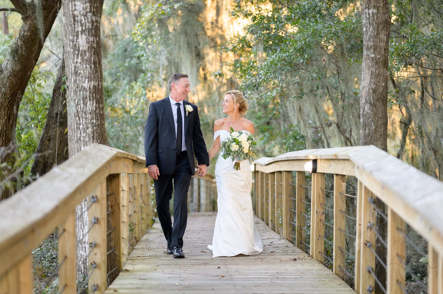 Bride and groom holding hands walking along a wooden boardwalk under mossy trees at Reserve Harbor Yacht Club