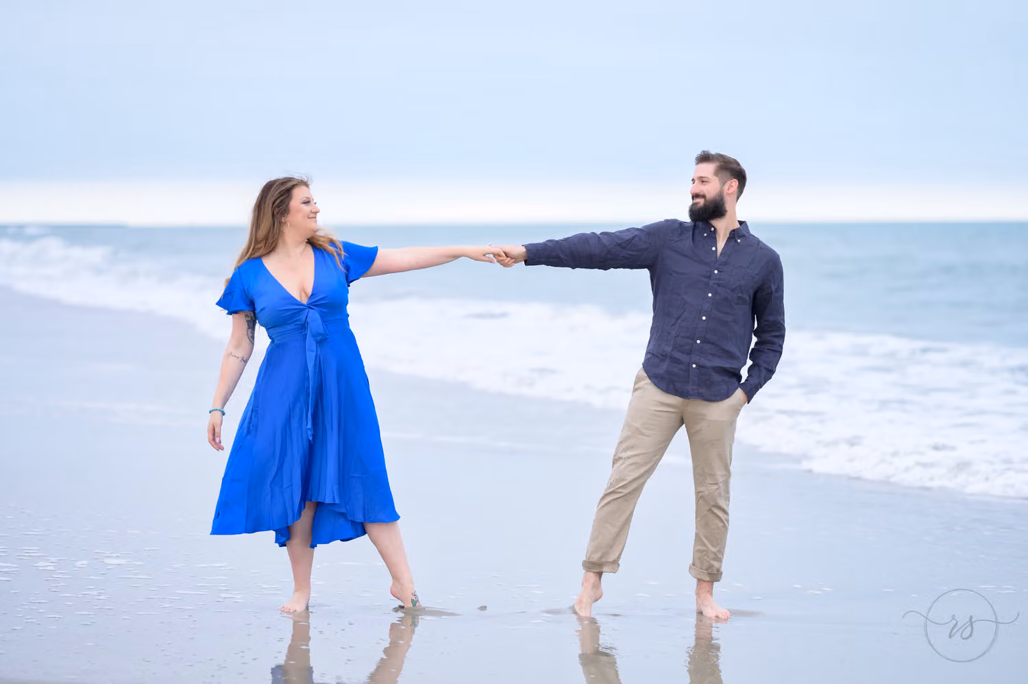 Woman in a bright blue dress holding hands with her fiancé as they lean apart on the beach in Myrtle Beach