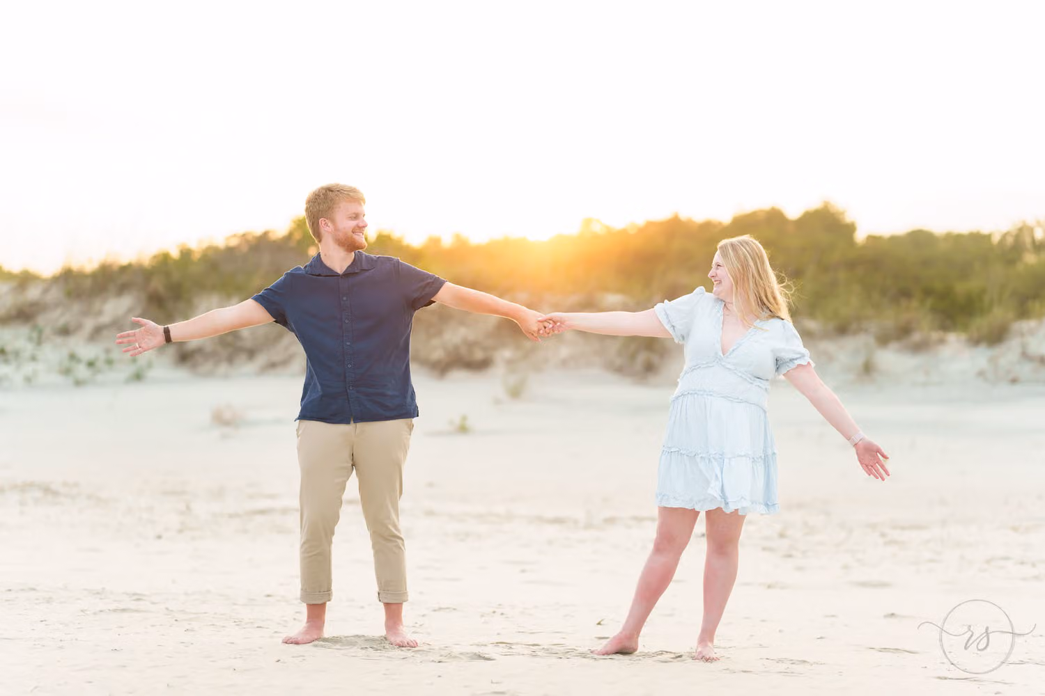Couple holding hands with arms stretched out on the sand as the sun sets behind the dunes at Huntington Beach State Park