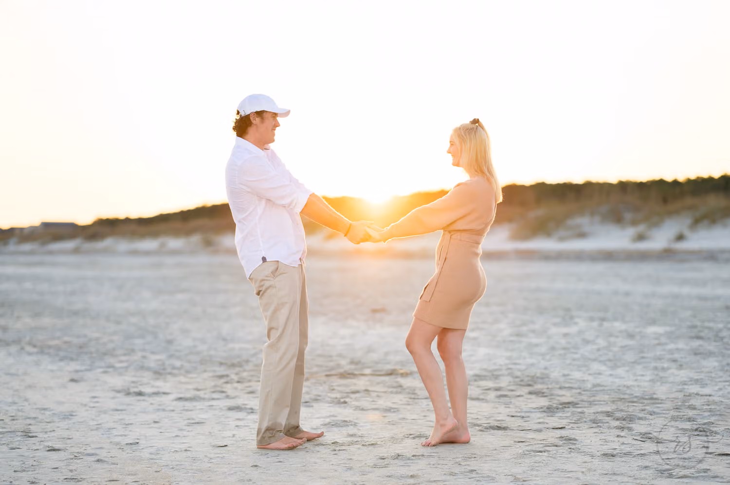 Couple holding hands at arm's length on the sand with the setting sun glowing between them at Huntington Beach State Park