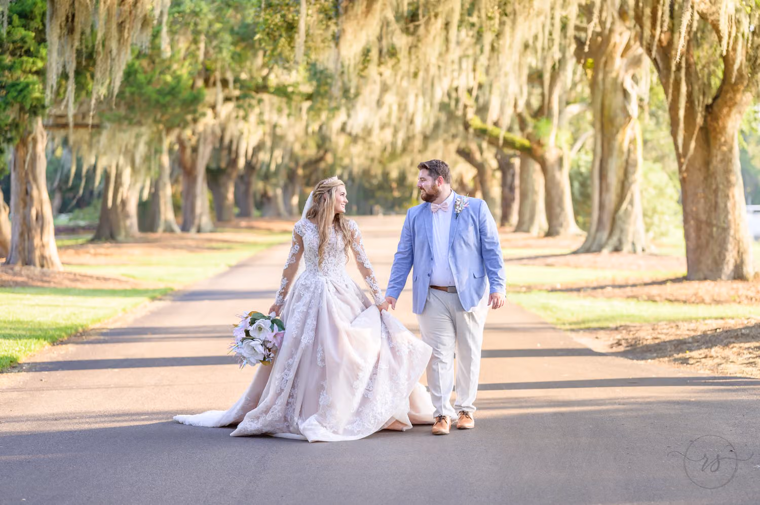 Bride and groom in a light blue suit walking hand in hand down a road under moss-draped oaks at Caledonia Golf & Fish Club