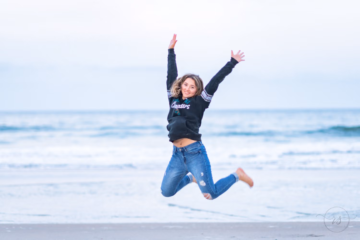 Senior in jeans and a black sweatshirt jumping high with her arms raised on the beach at Huntington Beach State Park