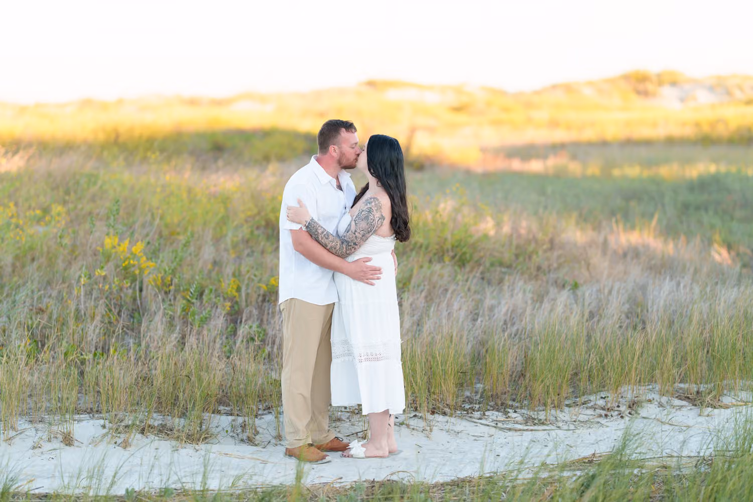 Couple kissing on a sandy path through tall dune grass and yellow wildflowers in warm evening light