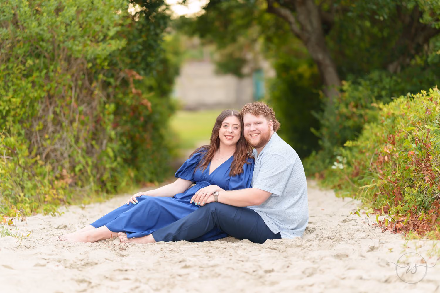 Newly engaged couple sitting close together on a sandy path under green trees at Huntington Beach State Park
