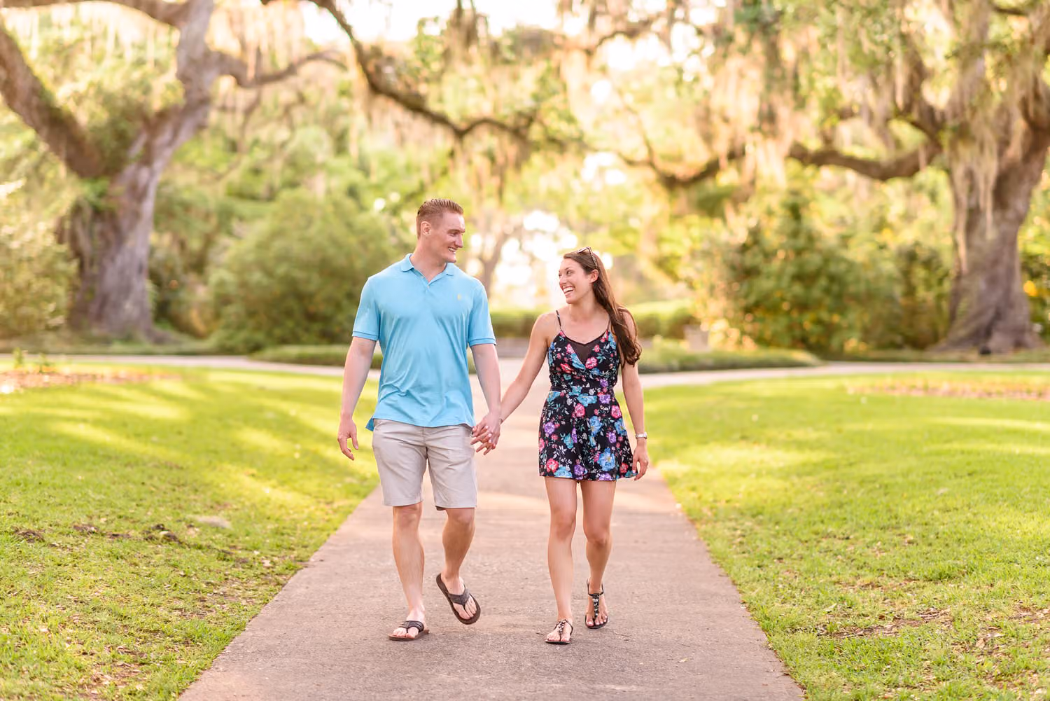 Couple holding hands as they walk down a paved path under oak trees with Spanish moss at Brookgreen Gardens