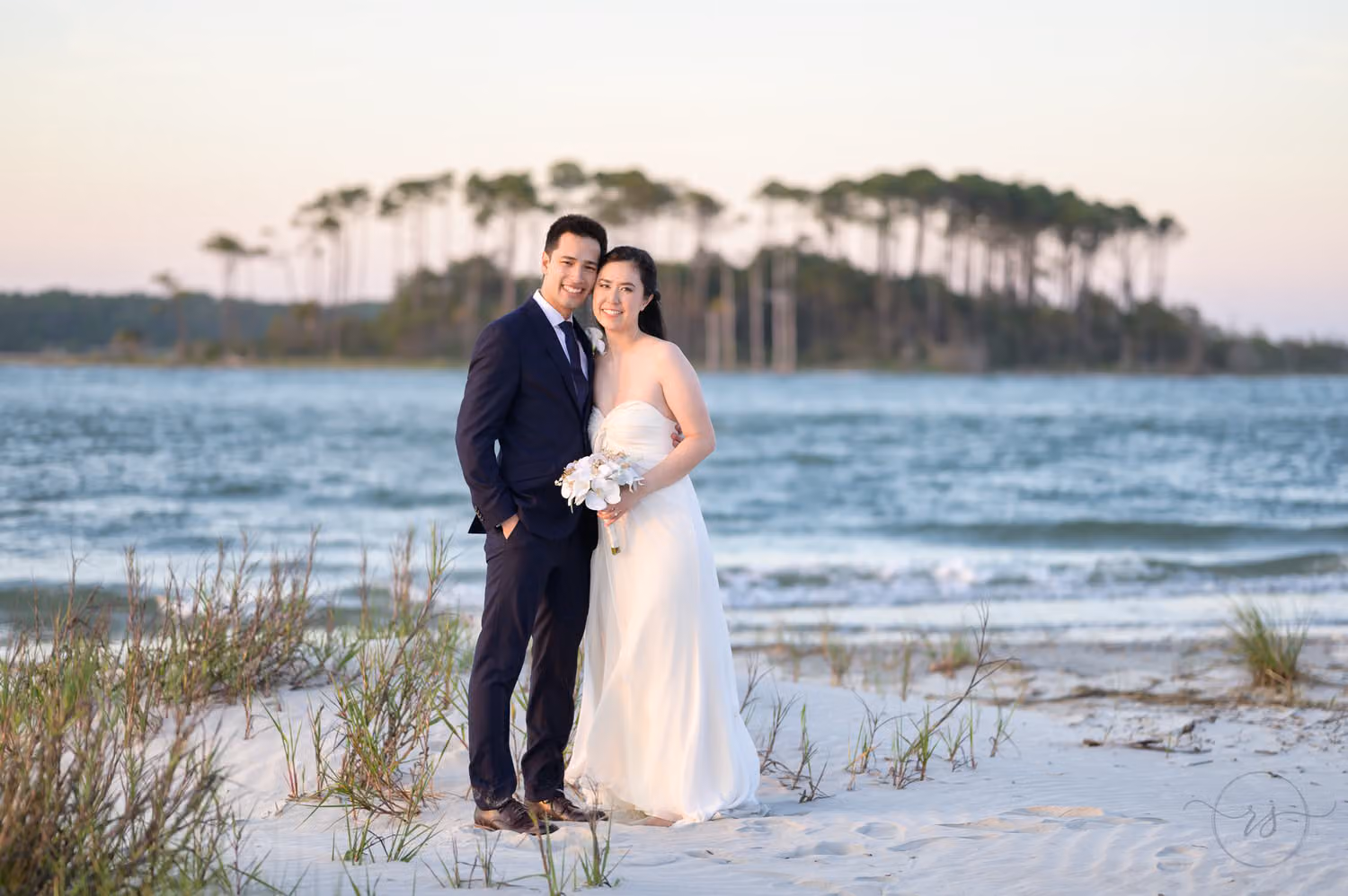 Bride and groom standing on the shore with a pine-covered island across the water at Cherry Grove Point in North Myrtle Beach