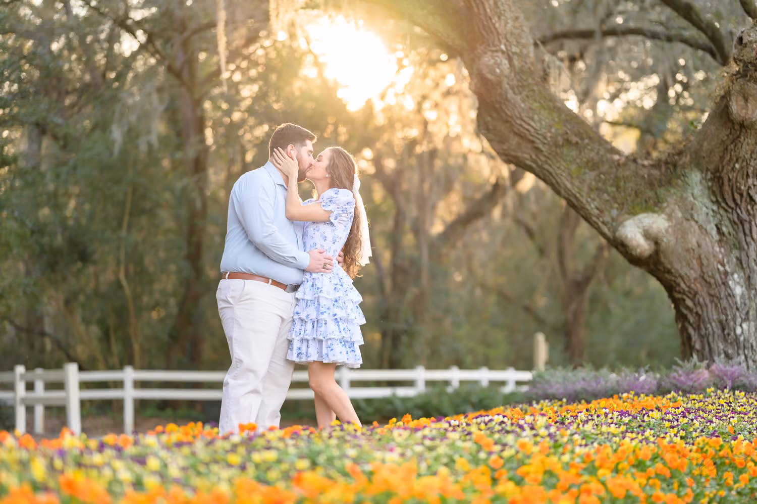 Couple kissing beside a white fence behind a bed of orange flowers in bloom under an oak at Caledonia Golf and Fish Club