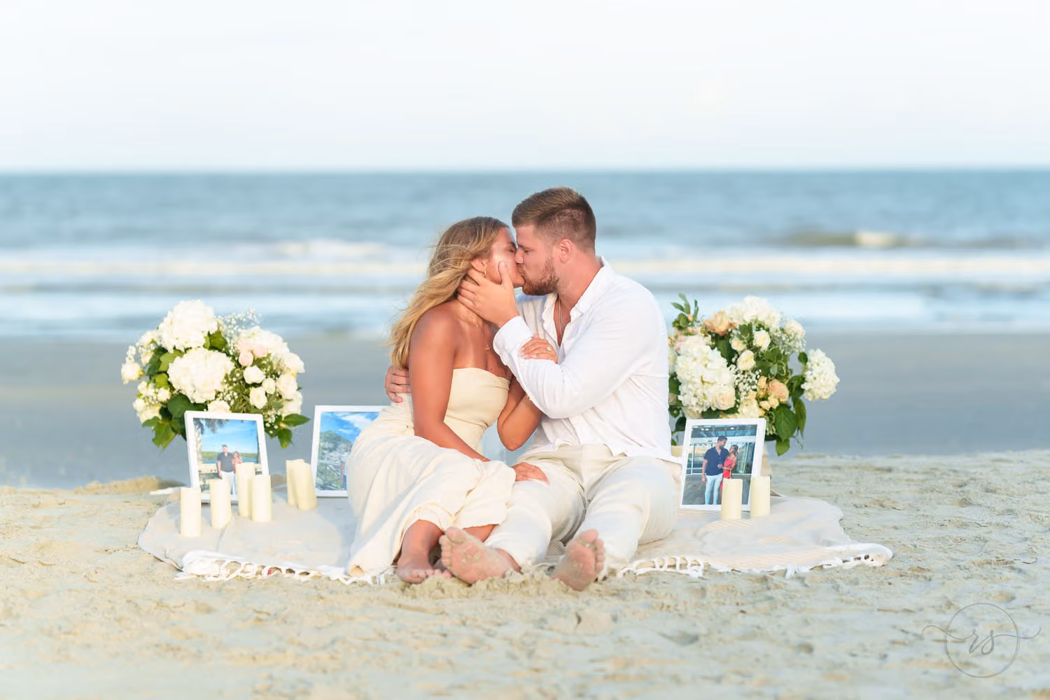 Engaged couple kissing on a blanket surrounded by white flowers, candles and framed photos on the beach in Pawleys Island