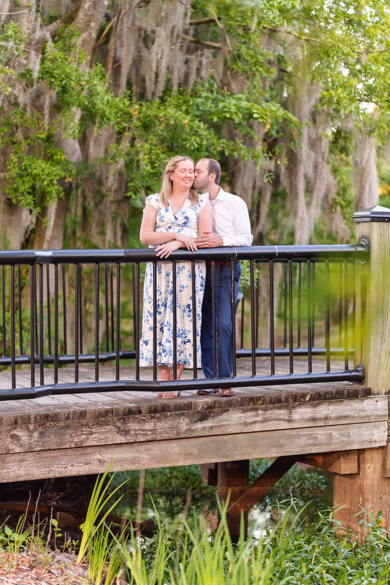 Couple kissing on a small bridge with a black railing under mossy trees along the Conway Riverwalk