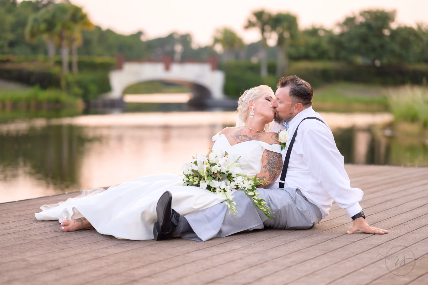 Bride and groom in suspenders kissing while sitting on a wooden dock by the pond and bridge at Wild Wing Plantation