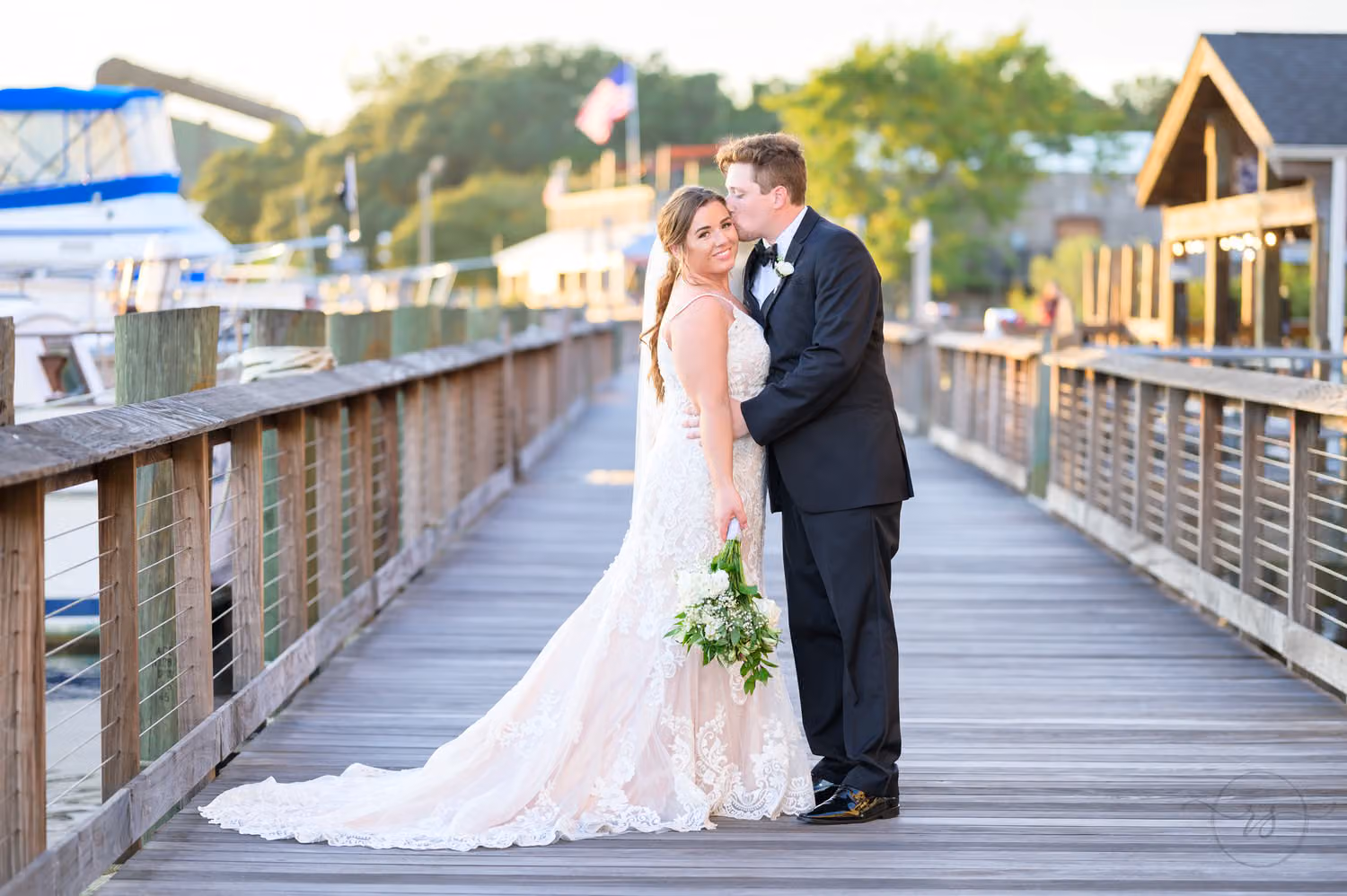 Groom kissing the bride on the cheek on the wooden boardwalk beside docked boats at the Georgetown Harborwalk