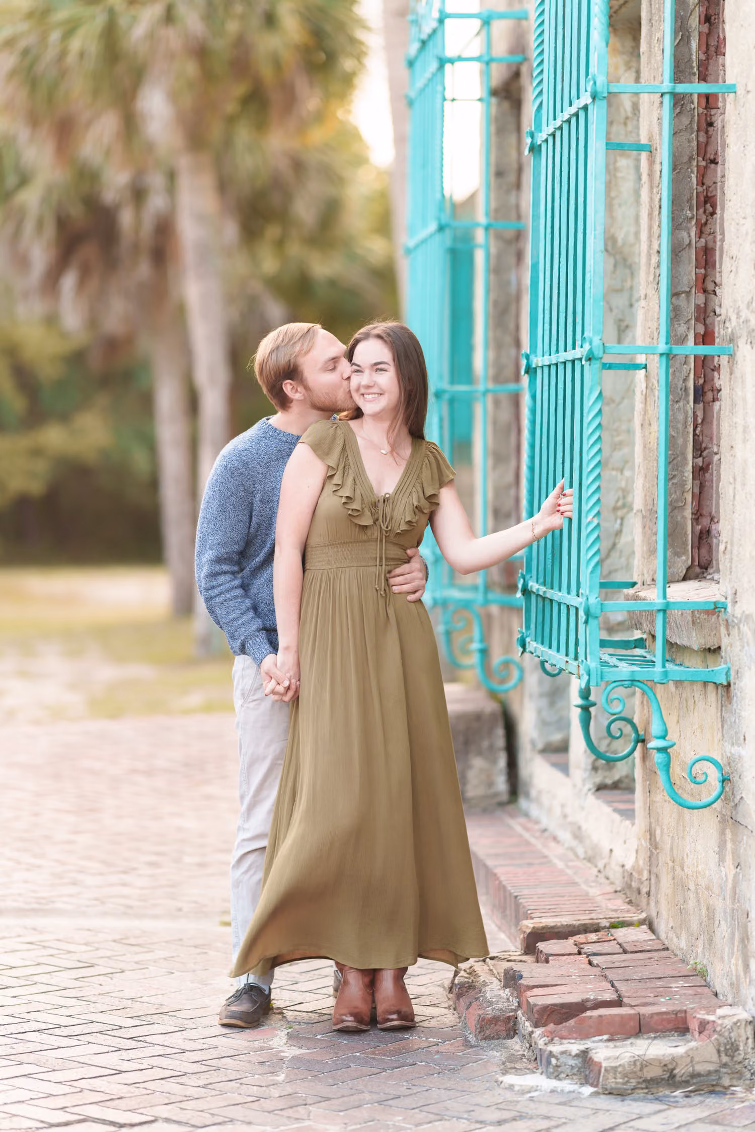 Man kissing his fiancée's cheek by the teal window grate of the Atalaya Castle at Huntington Beach State Park