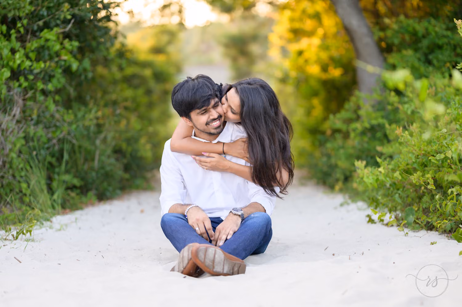 Woman hugging her fiancé from behind and kissing his cheek as they sit on a sandy path at Huntington Beach State Park