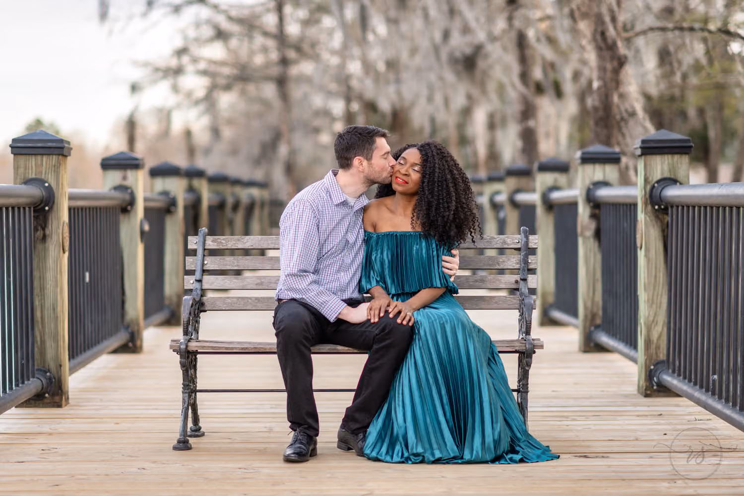 Man kissing his fiancée on the cheek as they sit on a bench on the boardwalk along the Conway Riverwalk