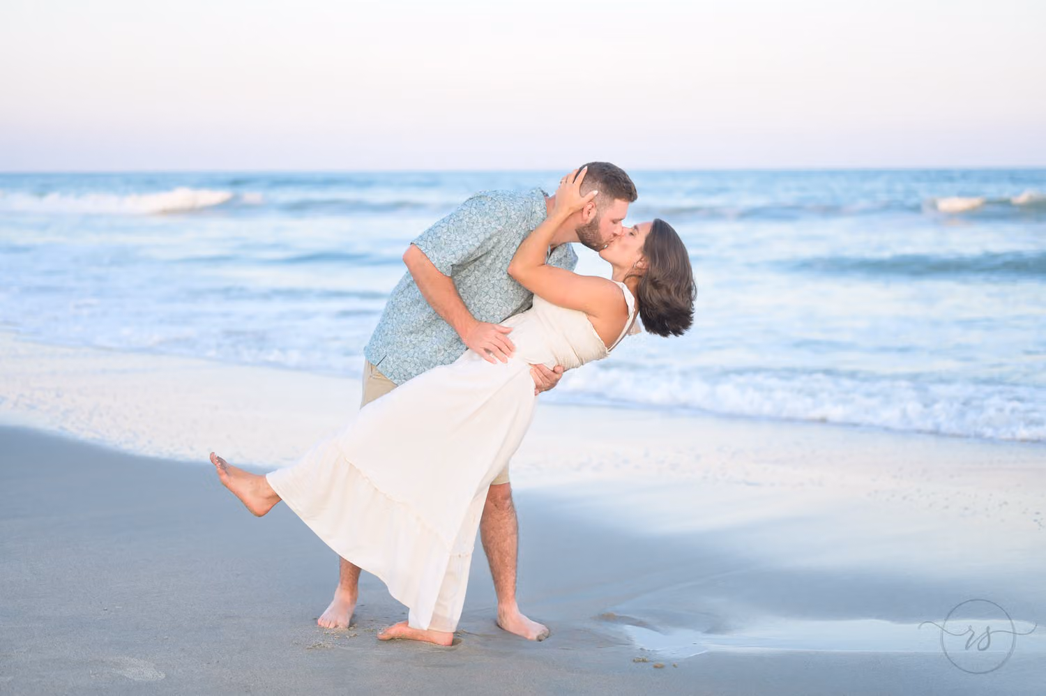 Man dipping his fiancée in a white sundress for a kiss in the shallow water at Huntington Beach State Park