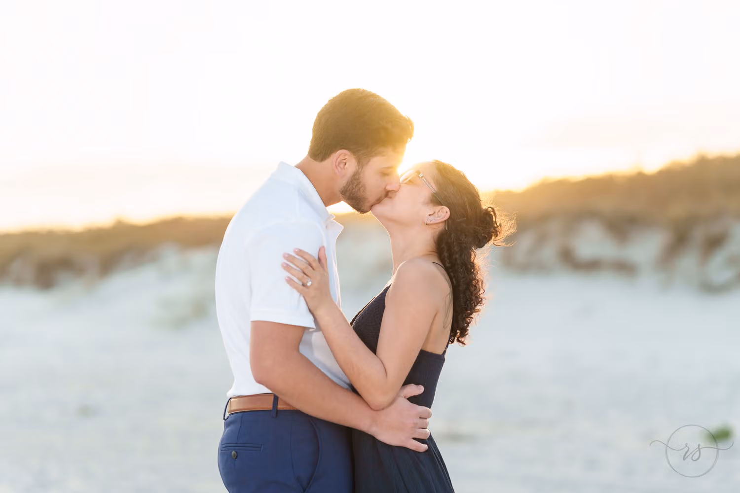 Newly engaged couple kissing in soft sunset light with the dunes behind them at Huntington Beach State Park