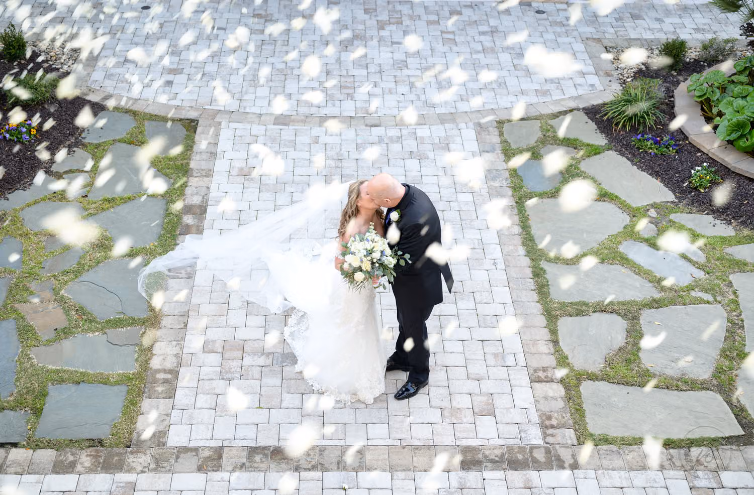 Overhead view of the bride and groom kissing on a stone path as white petals fall around them at 21 Main Events