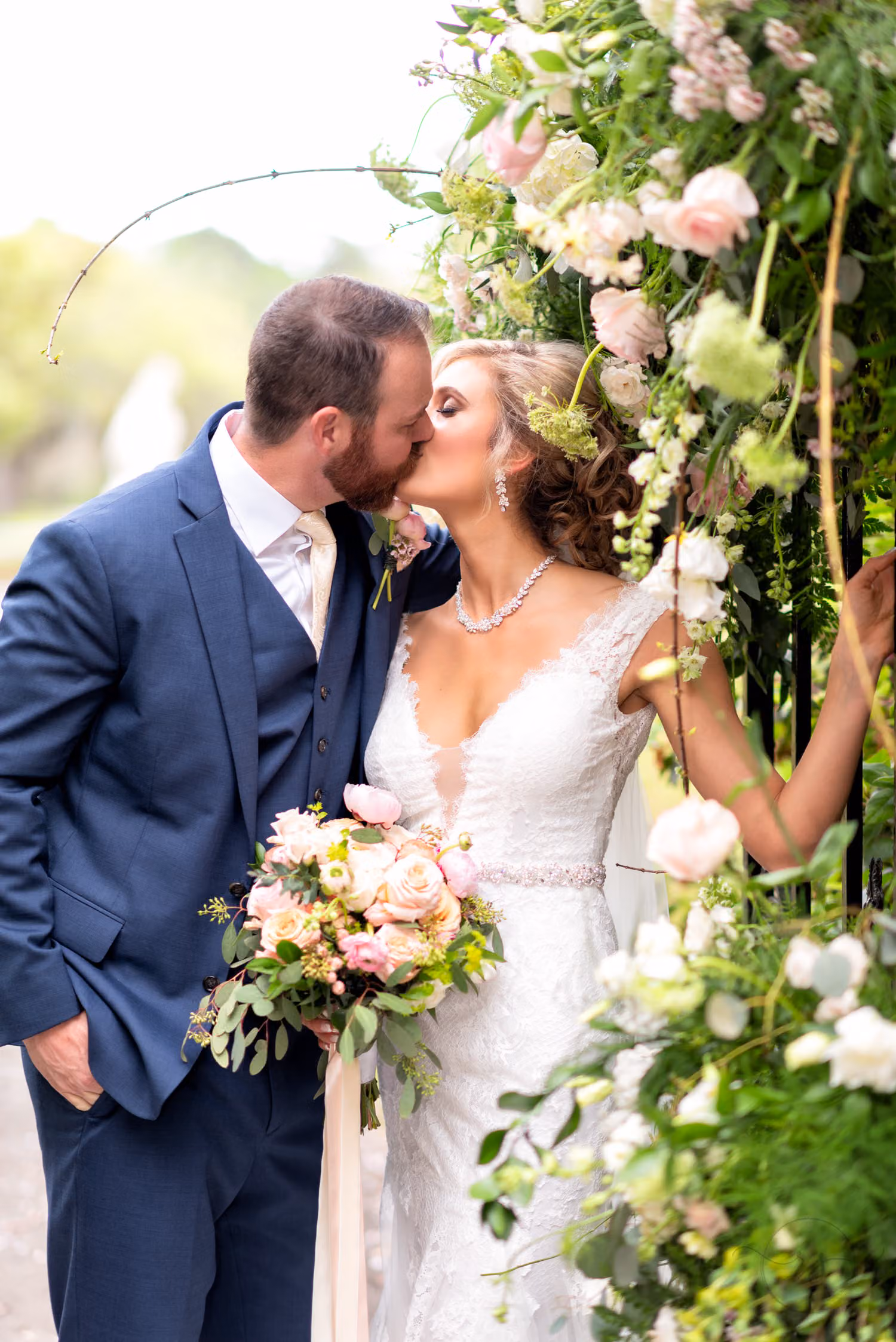 Bride and groom in a navy suit kissing beside a gate covered in pink and white flowers at Brookgreen Gardens