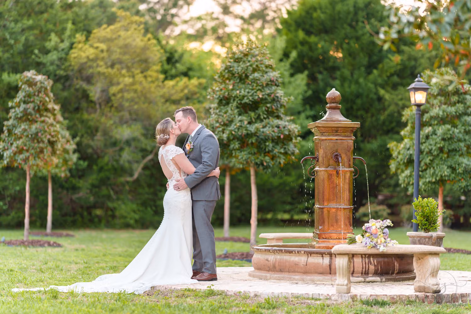 Bride and groom kissing beside a stone fountain and lamp post in the garden at Slate & Cypress Wedding and Event Venue