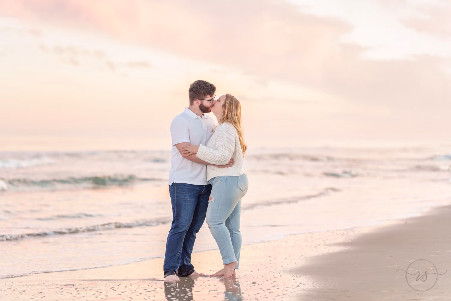 Couple kissing at the water's edge under soft pink cotton candy clouds at Huntington Beach State Park