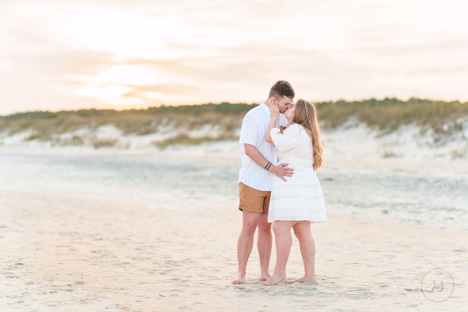 Couple kissing on the wide beach with sandy dunes behind them in soft evening light at Huntington Beach State Park