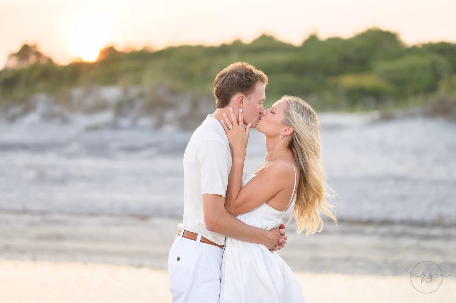 Couple in white kissing on the beach with the dunes and setting sun behind them at Huntington Beach State Park