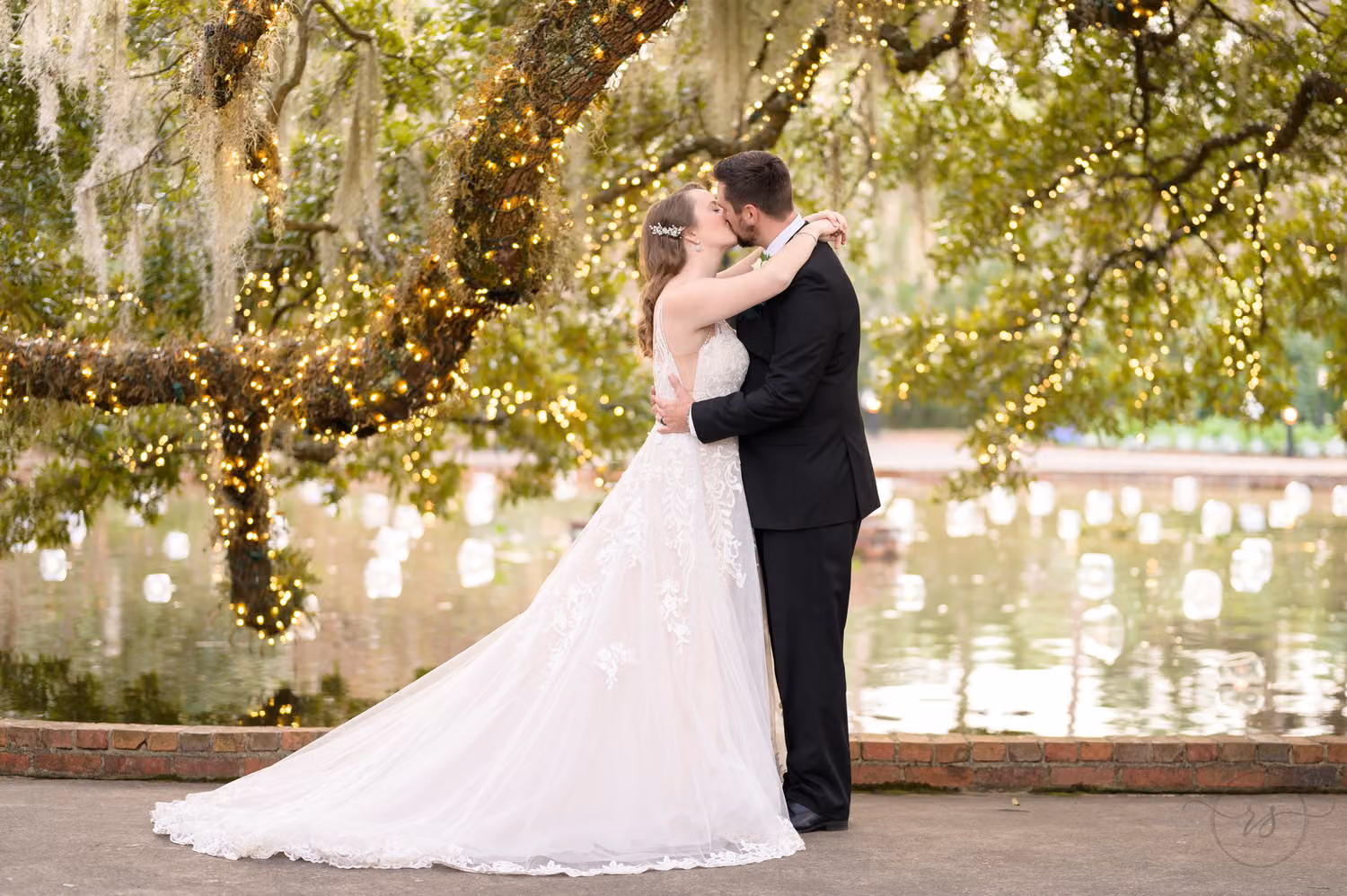 Bride and groom kissing by the reflecting pool with string lights and candles glowing in the oaks at Brookgreen Gardens