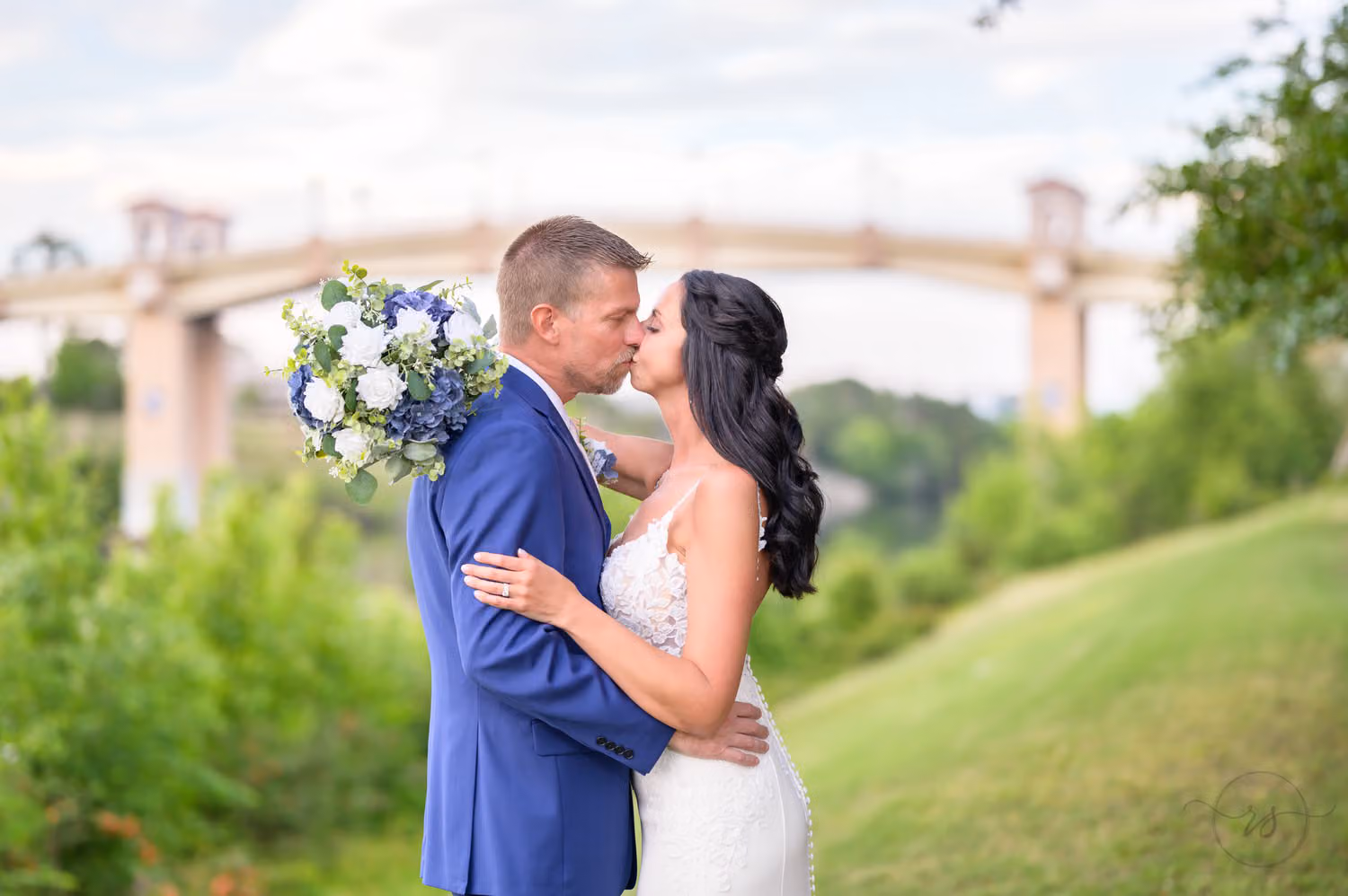 Bride and groom in a blue suit kissing on a green hill with the Grande Dunes bridge behind them at The Marina Inn