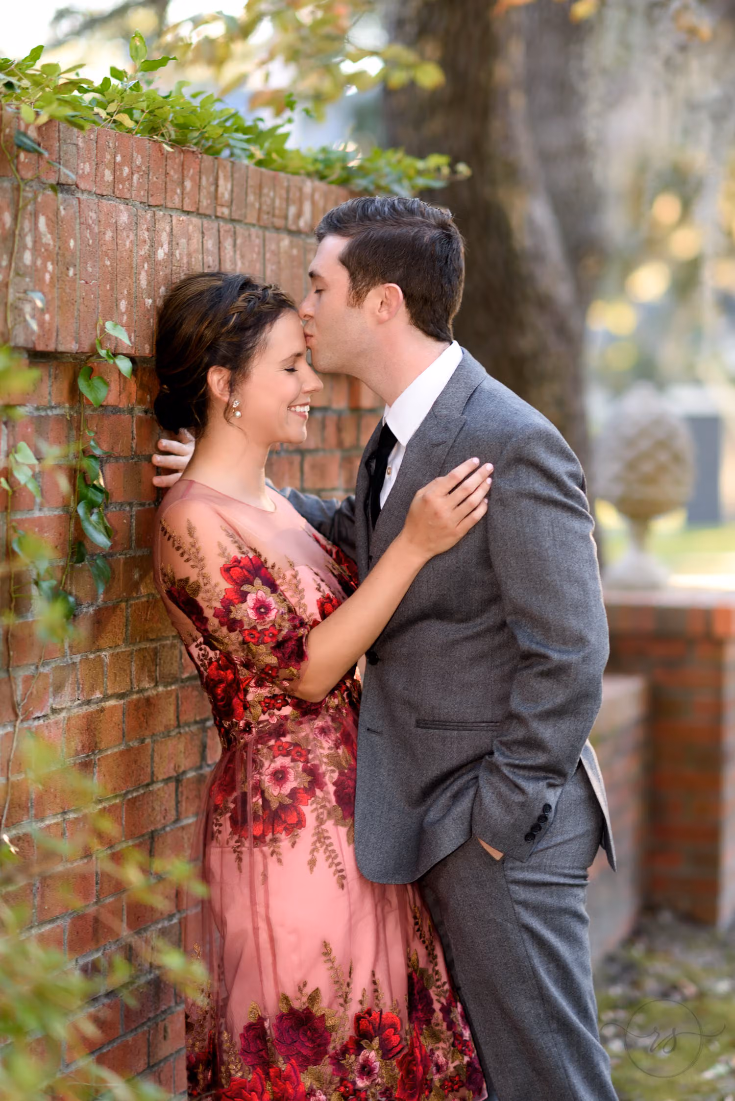 Man kissing his fiancée's head as she leans on an old brick wall in a red floral dress on Oak Street in Conway