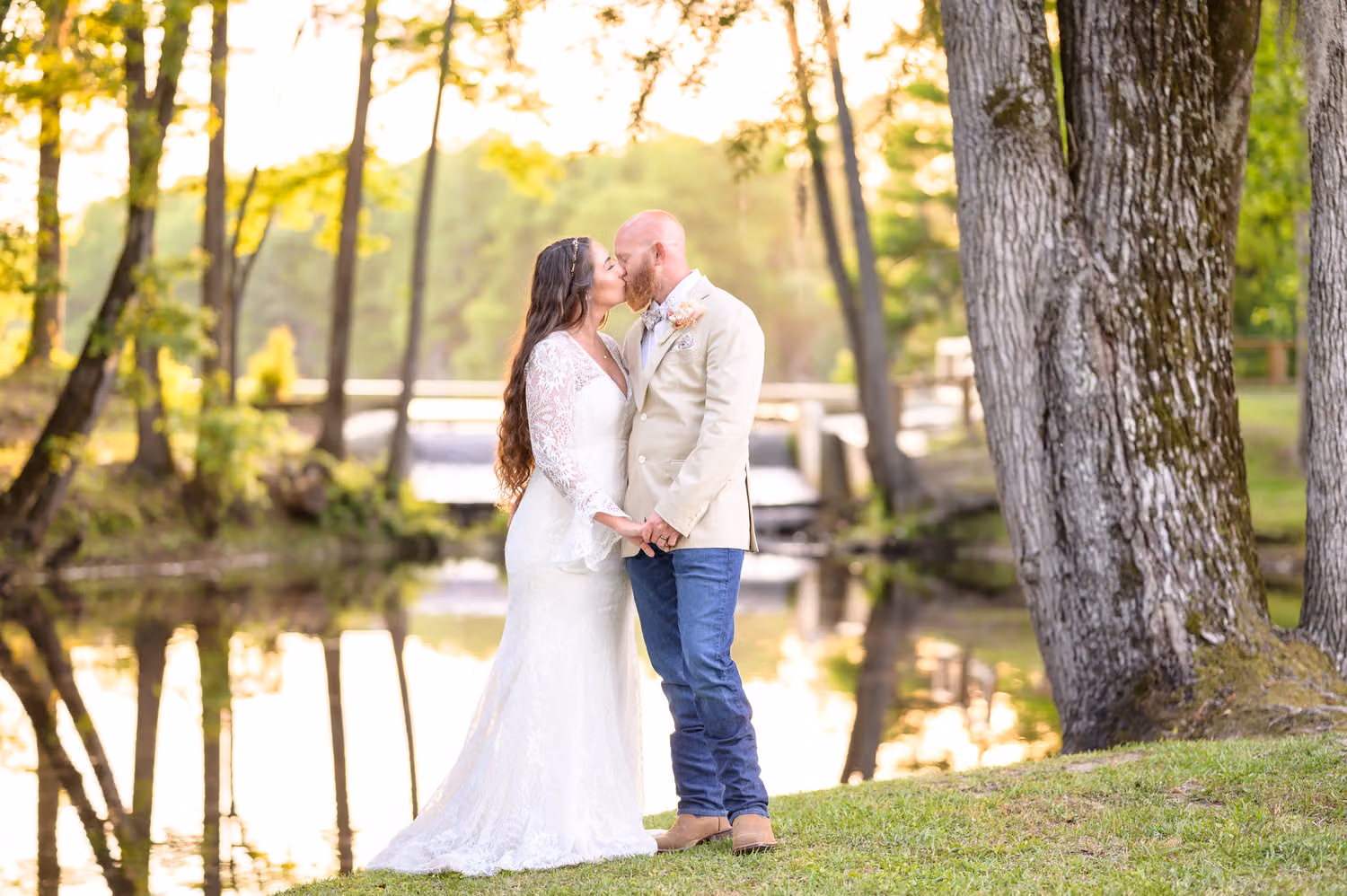 Bride and groom in a tan jacket and jeans kissing beside a big tree at the edge of the lake at The Spillway