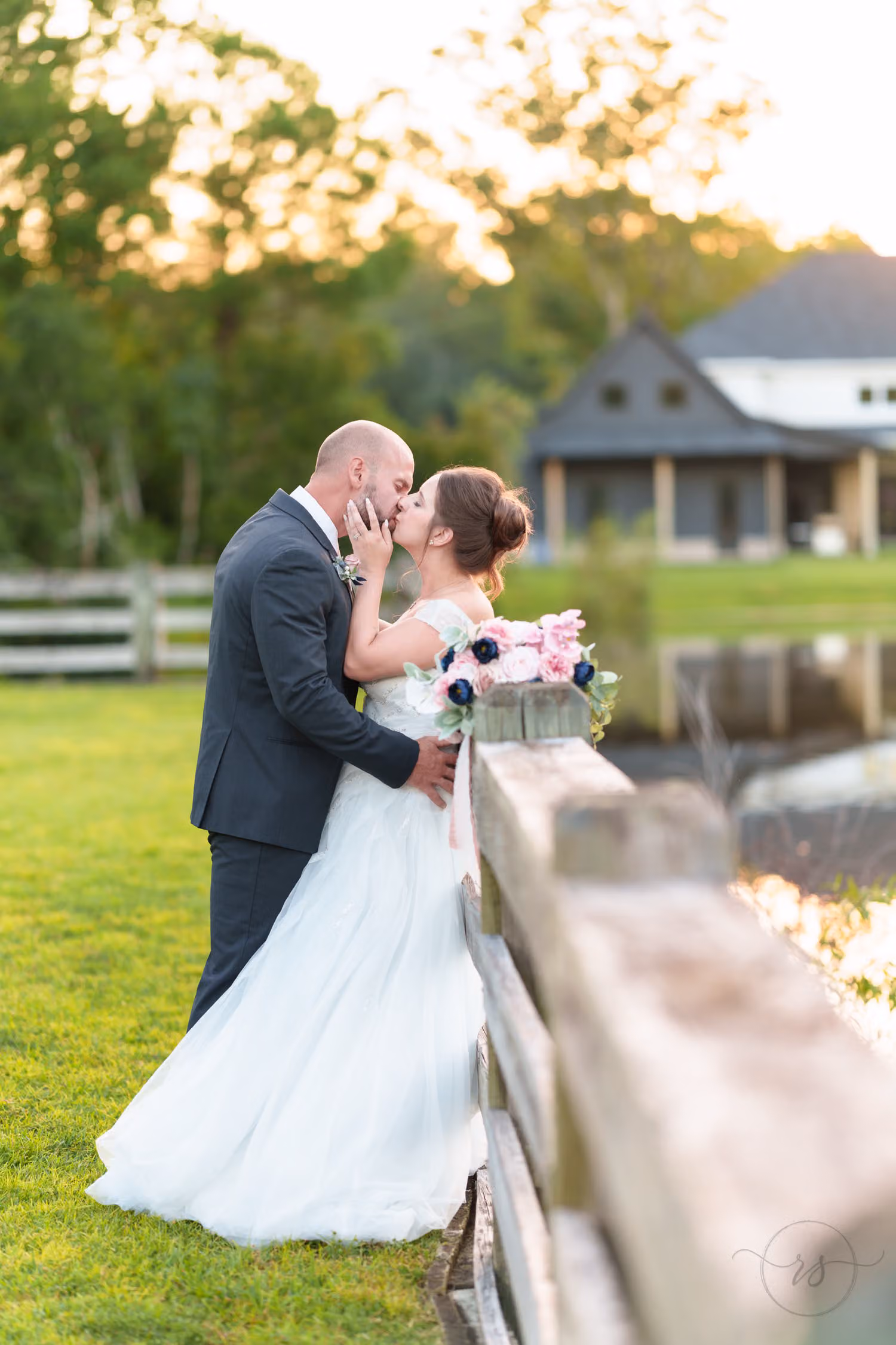 Bride and groom kissing as they lean on a wooden fence by a pond in golden light at The Pavilion at Pepper Plantation