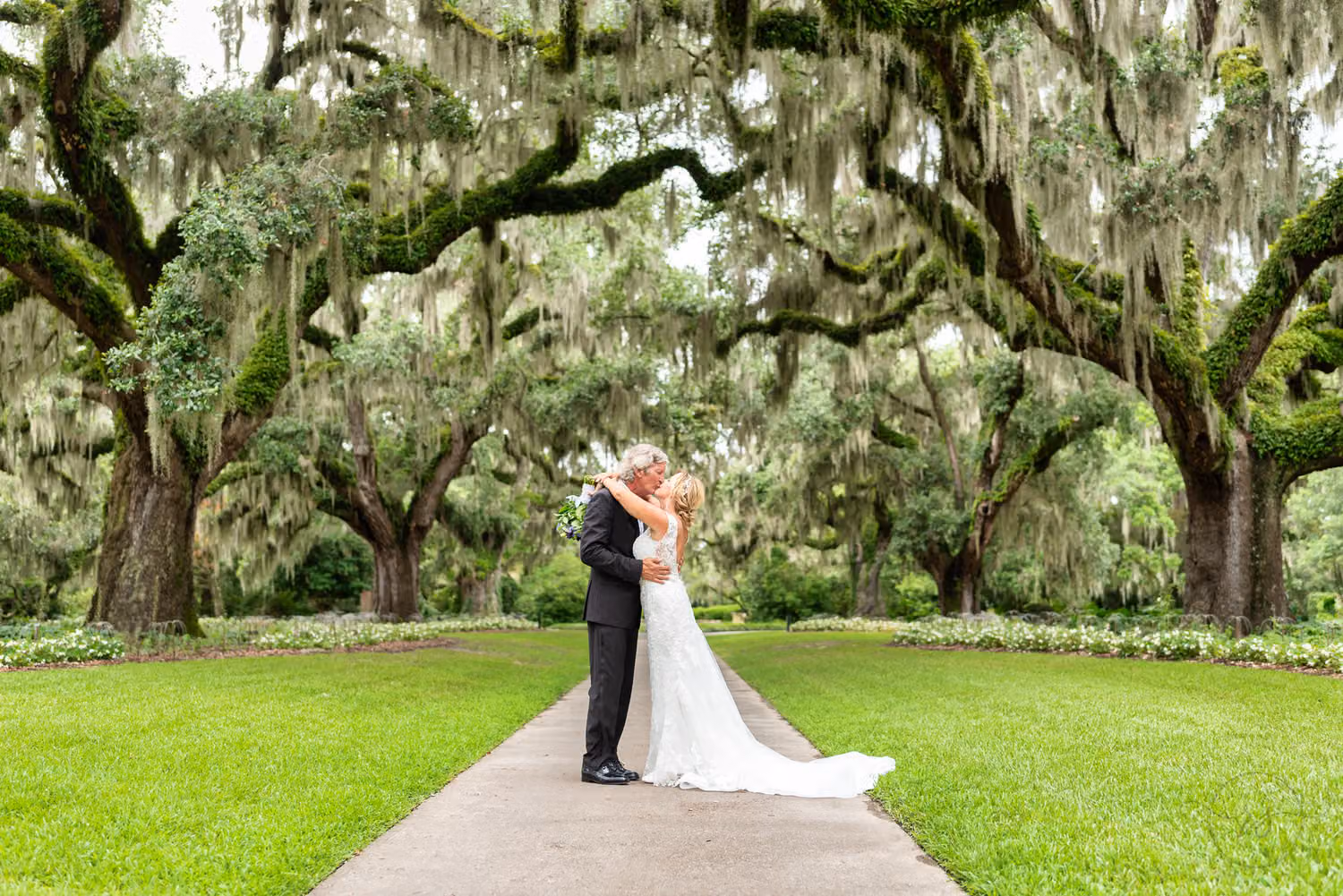 Bride and groom kissing on a paved path beneath huge moss-draped live oaks on the Live Oak Allee at Brookgreen Gardens