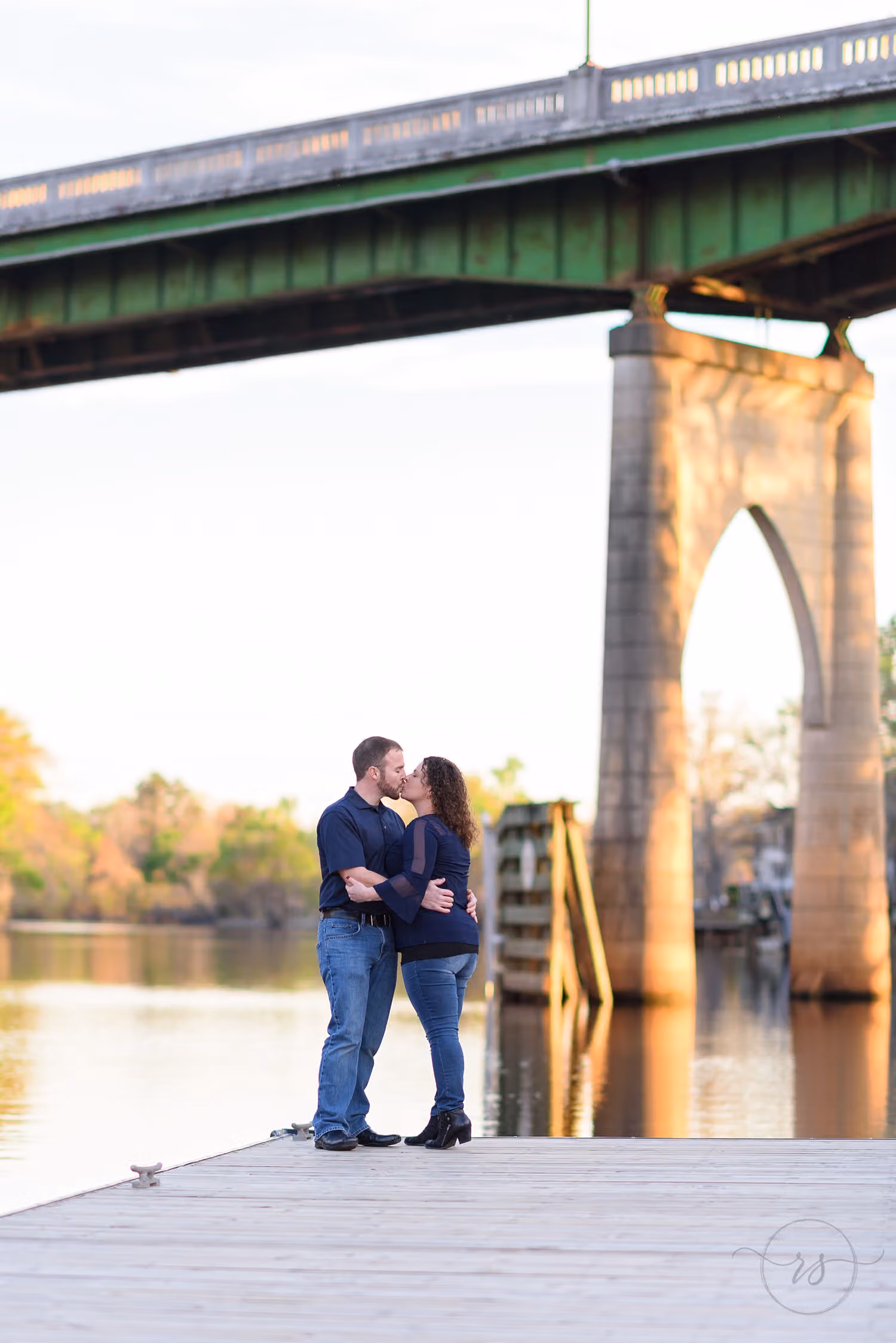 Couple kissing on a dock by the river beneath a green bridge with stone arches along the Conway Riverwalk