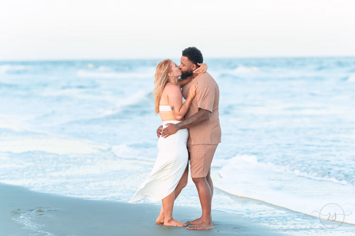 Couple kissing at the water's edge as small waves wash over their feet at Huntington Beach State Park