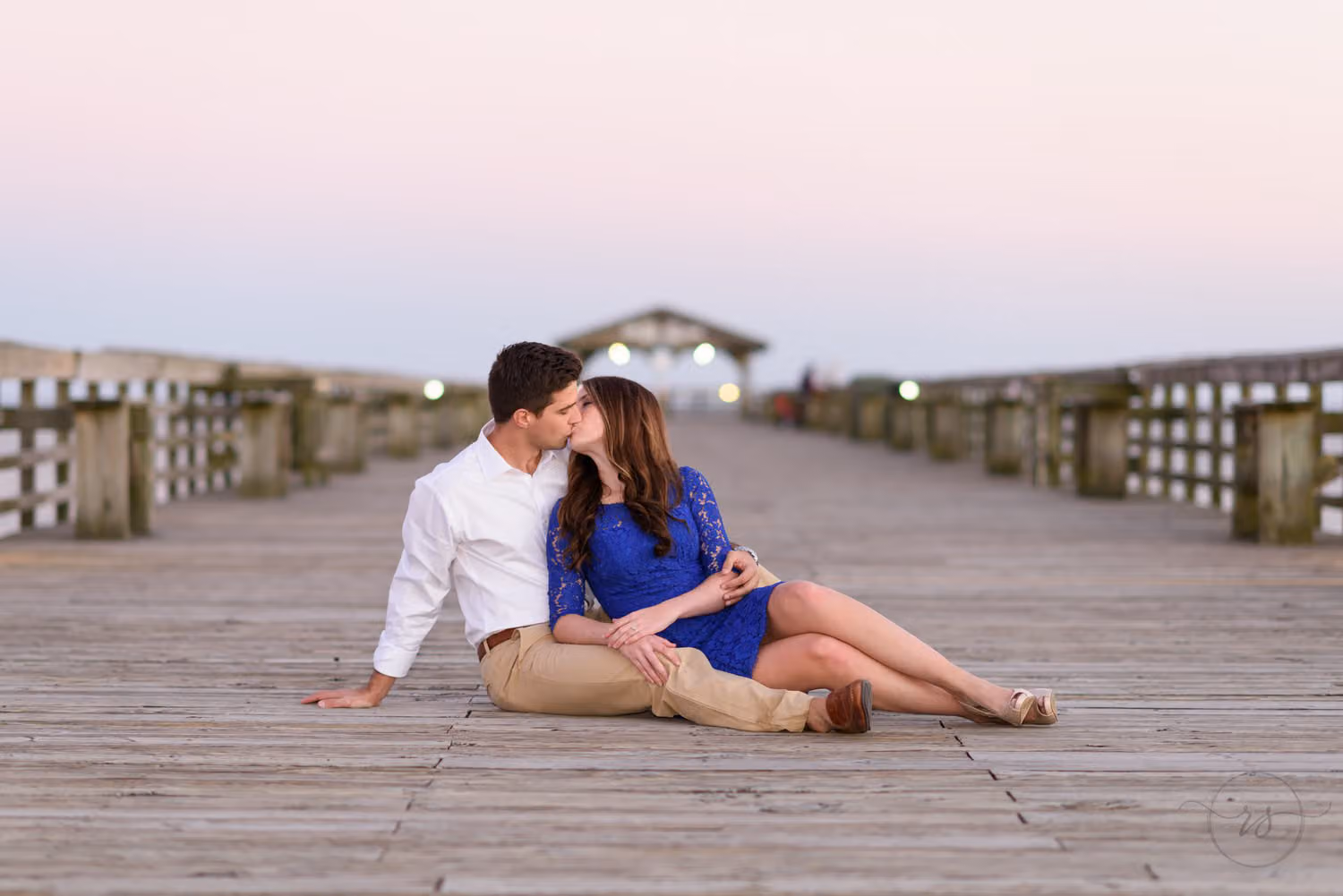 Couple sitting on the wooden pier leaning in for a kiss under a soft pink sky at Myrtle Beach State Park