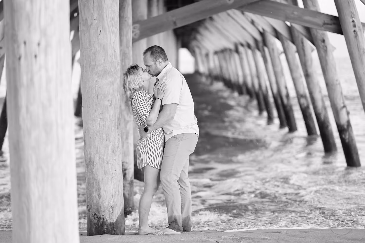 Black and white photo of a couple kissing between the wooden pilings under the pier at Myrtle Beach State Park