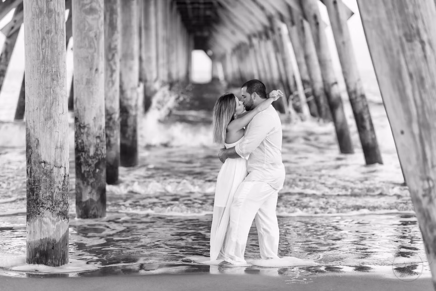 Black and white photo of a couple kissing under the pier as waves splash around the pilings at Myrtle Beach State Park