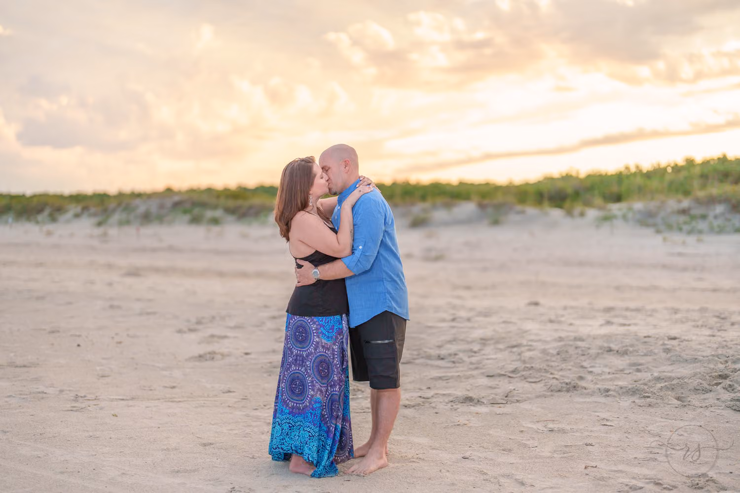 Couple kissing on the sand in front of the dunes under a soft sunset sky at Huntington Beach State Park
