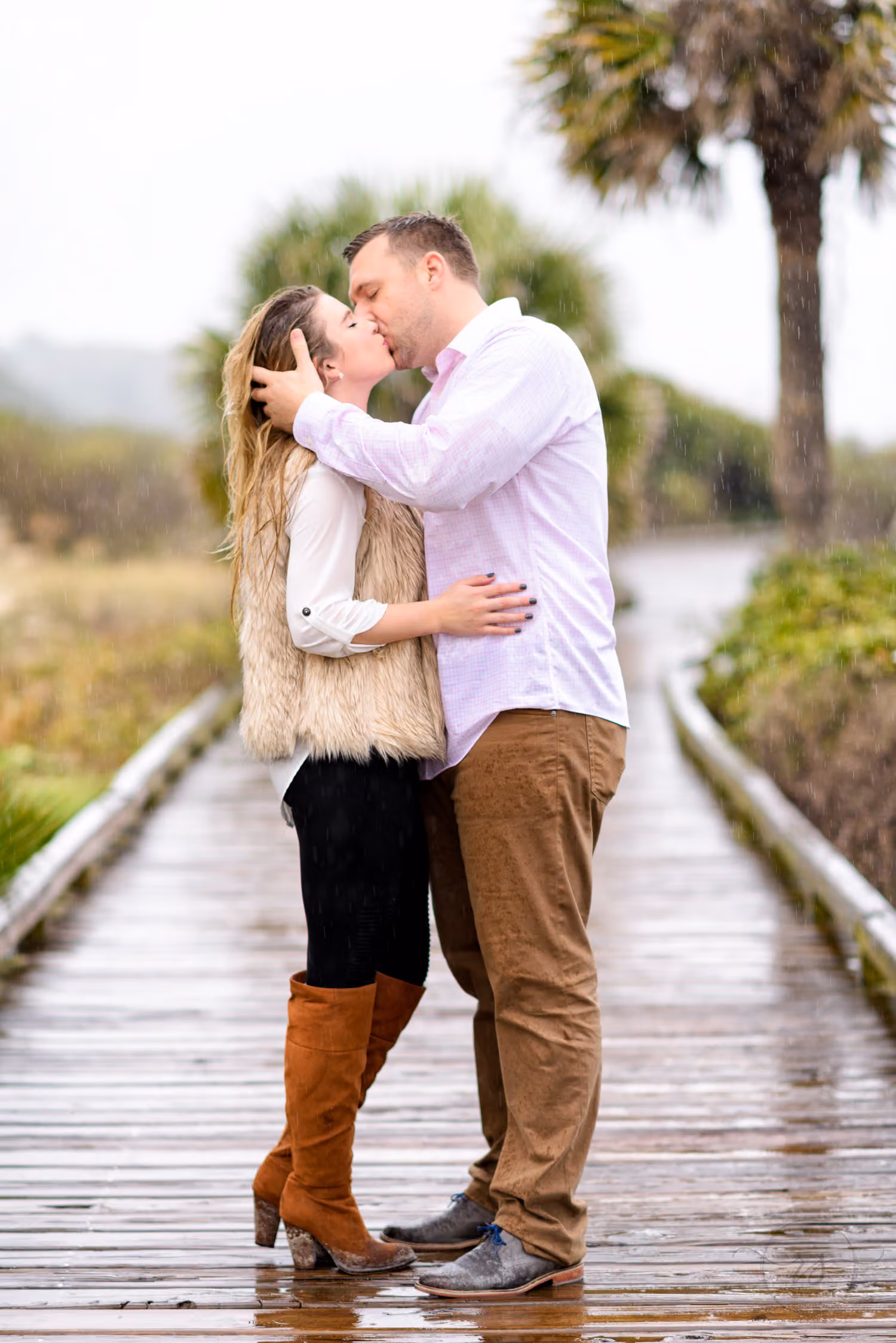 Couple kissing on a wet wooden boardwalk lined with palmetto trees on a rainy day in Pawleys Island