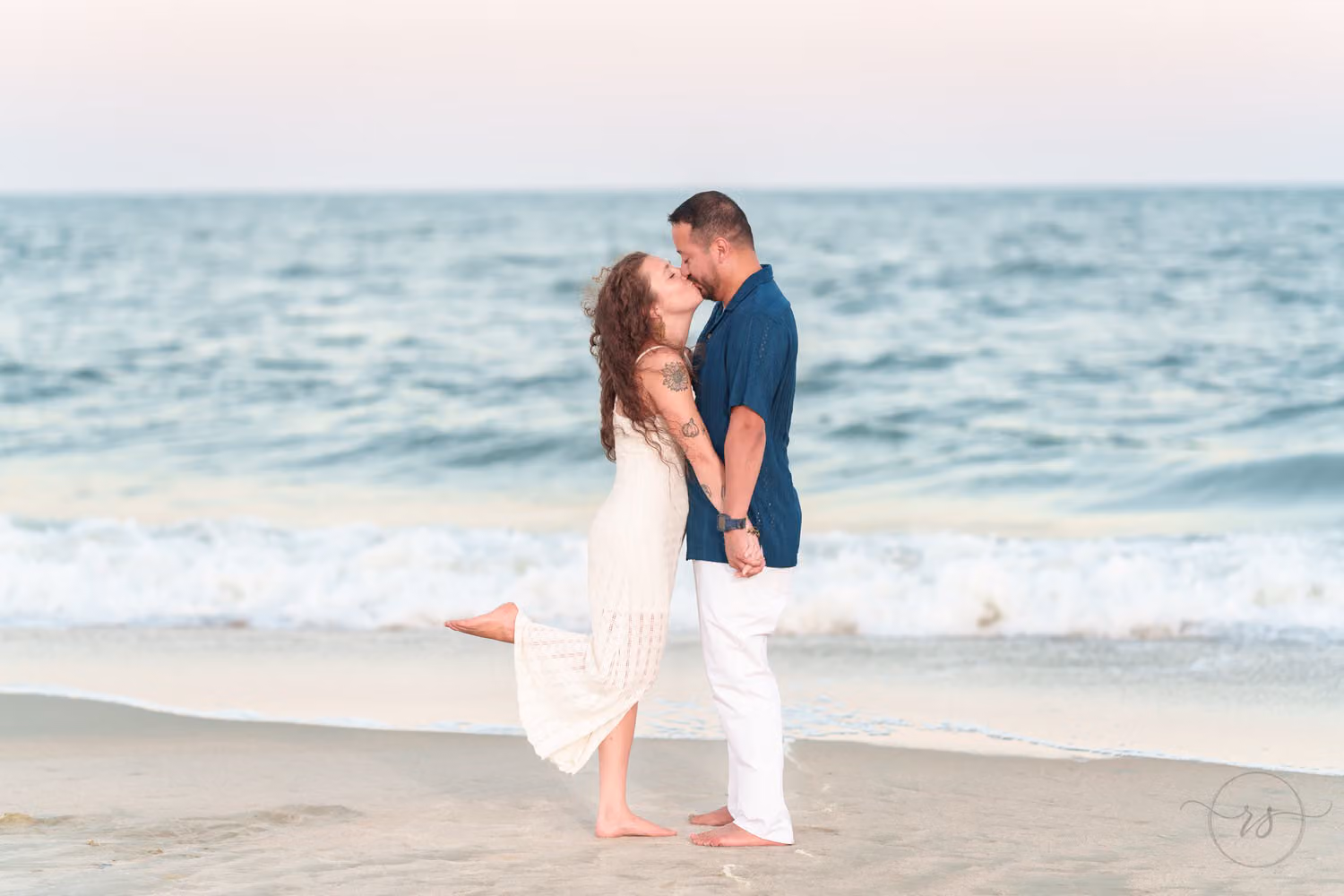 Couple kissing at the water's edge as she lifts one foot in the shallow waves at Huntington Beach State Park
