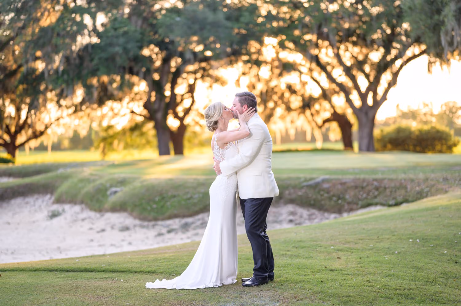 Bride and groom in a white jacket kissing beside a sand bunker on the golf course at sunset at Caledonia Golf & Fish Club