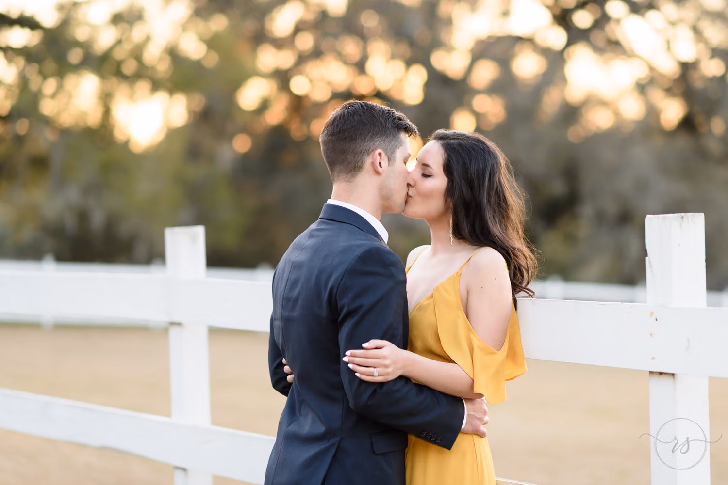 Couple sharing a kiss by a white fence with golden sunset light glowing through the trees at Mansfield Plantation