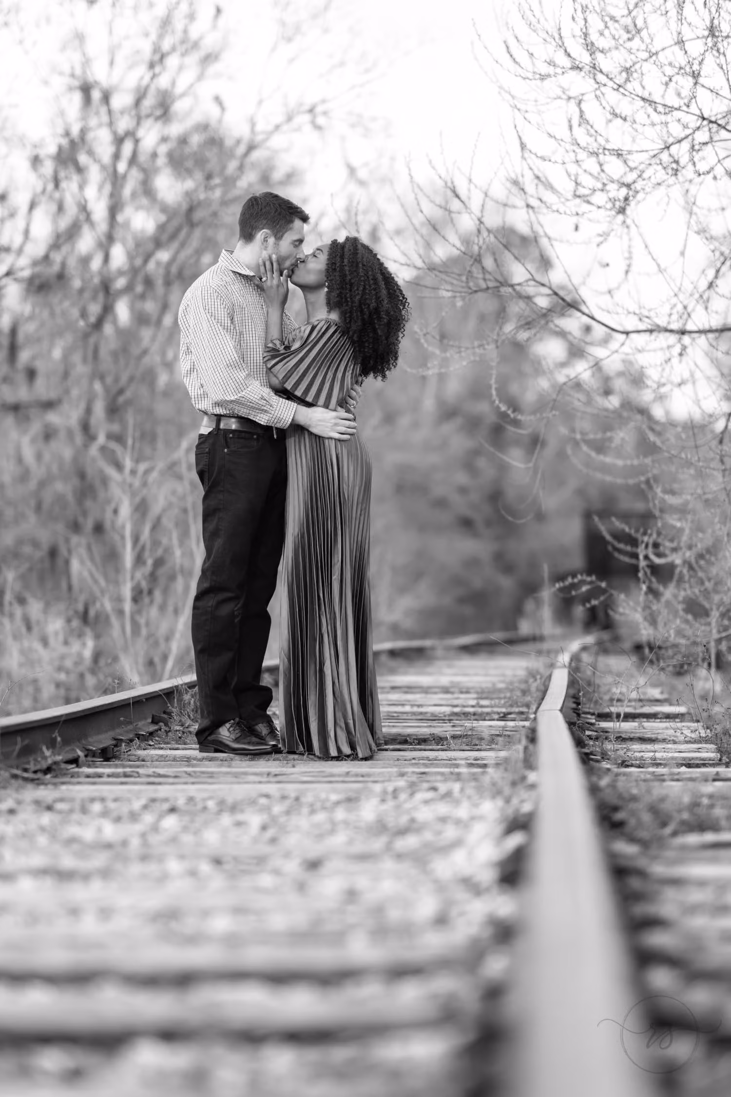 Black and white photo of a couple kissing on old train tracks lined with bare trees by the Conway Riverwalk