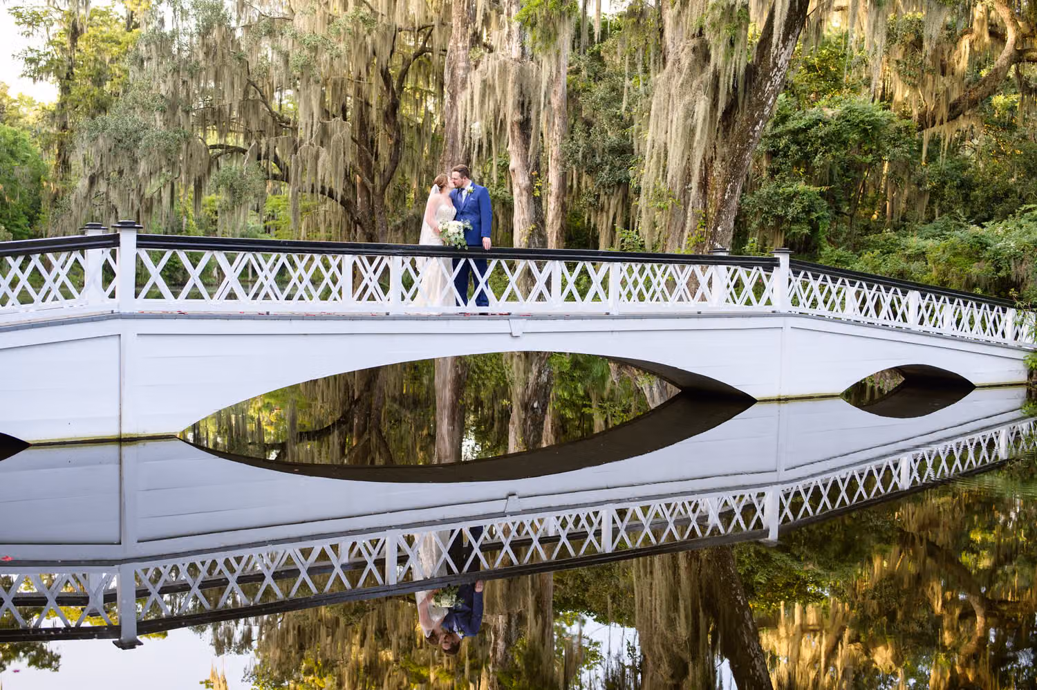 Bride and groom kissing on the white arched bridge reflected in the still water at Magnolia Plantation in Charleston