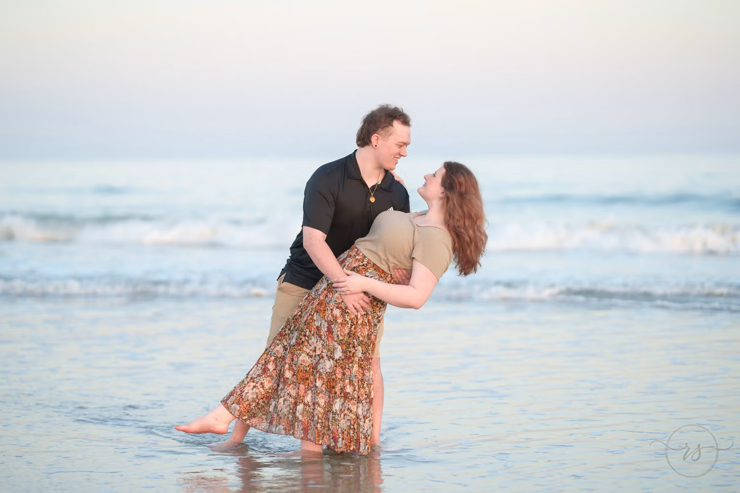 Man dipping his fiancée in a long floral skirt as she laughs in the shallow water at Huntington Beach State Park